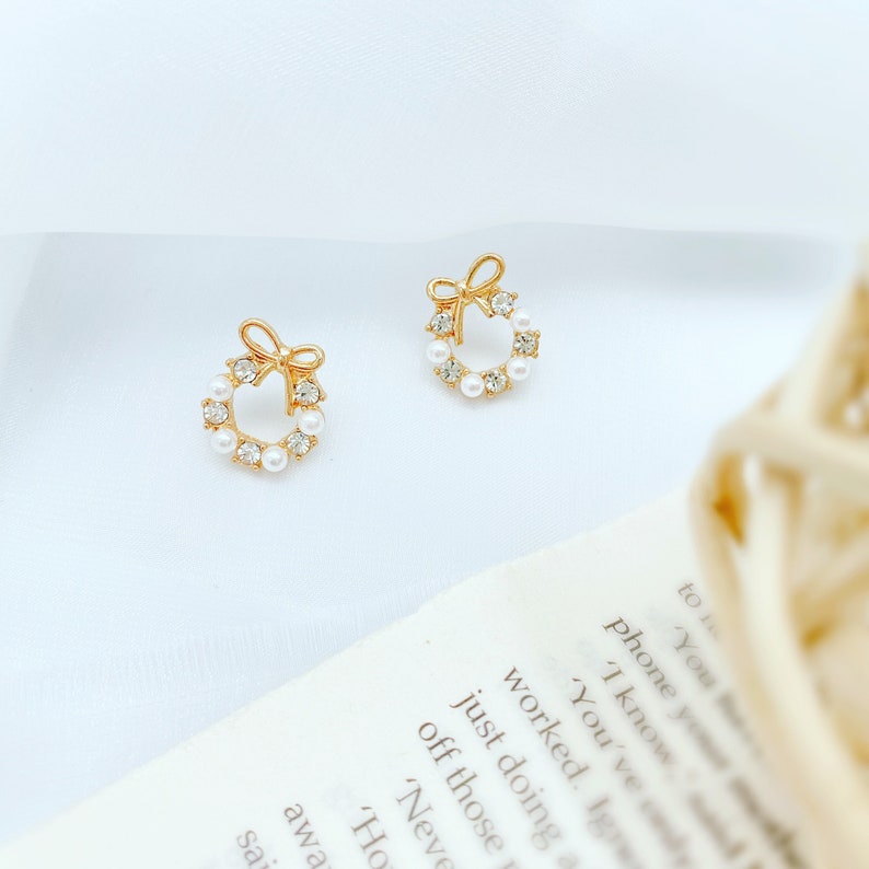 May include: A pair of gold-tone earrings with a small pearl and rhinestone wreath design. Each earring features a small bow at the top.