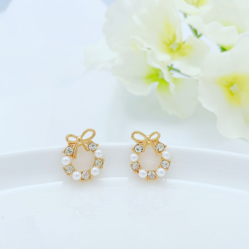 May include: A pair of gold tone earrings with a pearl and rhinestone wreath design. Each earring features a small bow at the top.