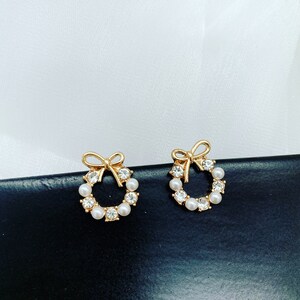 May include: A pair of gold-tone earrings with a wreath design. The wreath is adorned with white pearls and clear rhinestones. The earrings have a bow detail at the top.