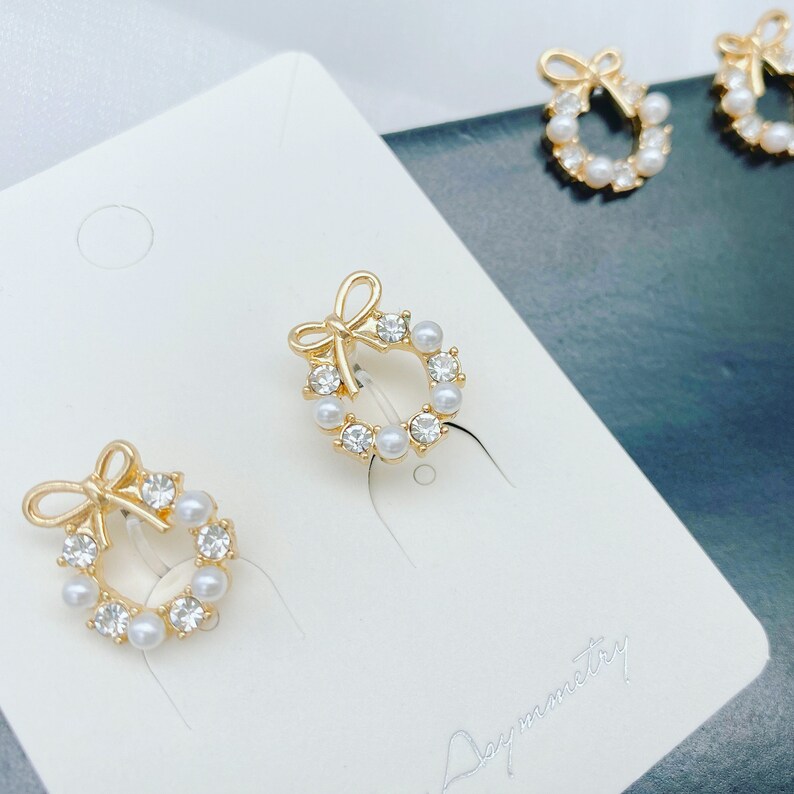 May include: A pair of gold-tone earrings with a wreath design. The wreath is adorned with small white pearls and rhinestones. The earrings have a bow at the top.