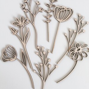 May include: Set of six laser-cut wooden floral shapes, including a tulip, a daisy, a poppy, and three different types of leafy stems. The shapes are all in a light brown color and have a simple, minimalist design.