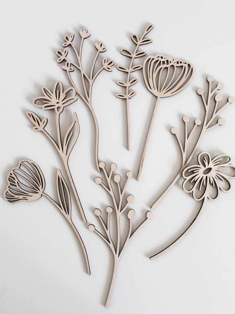 May include: Set of six laser-cut wooden floral shapes, including a tulip, a daisy, a poppy, and three different types of leafy stems. The shapes are all in a light brown color and have a simple, minimalist design.