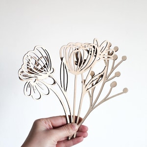 May include: A set of three wooden flower cutouts, featuring a large flower with a rounded center, a smaller flower with a more open center, and a delicate stem with small round buds. The cutouts are made of light brown wood and are held in a hand.