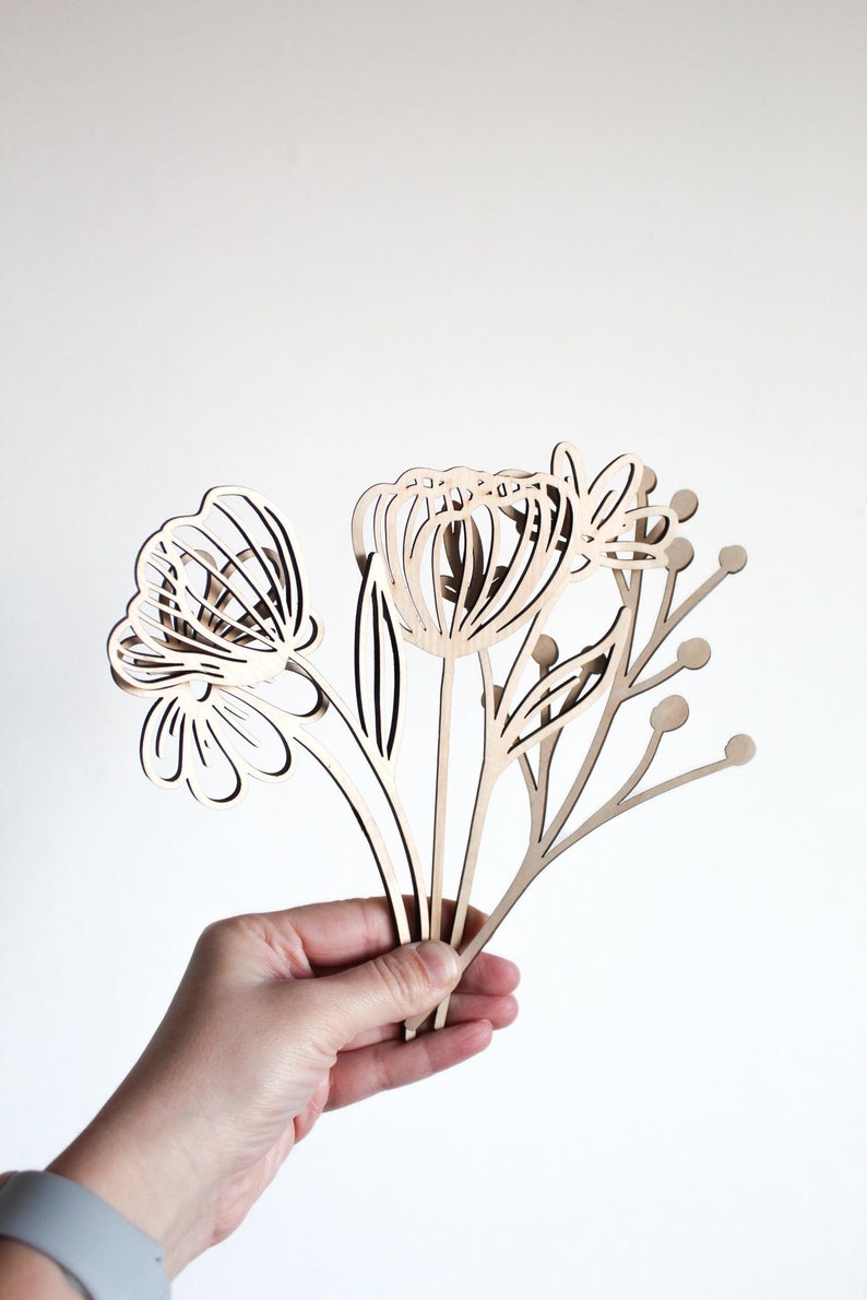 May include: A set of three wooden flower cutouts, featuring a large flower with a rounded center, a smaller flower with a more open center, and a delicate stem with small round buds. The cutouts are made of light brown wood and are held in a hand.