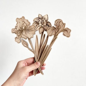 May include: A set of three wooden iris flower cutouts in a light brown color. The flowers are arranged in a bouquet and are held in a hand.