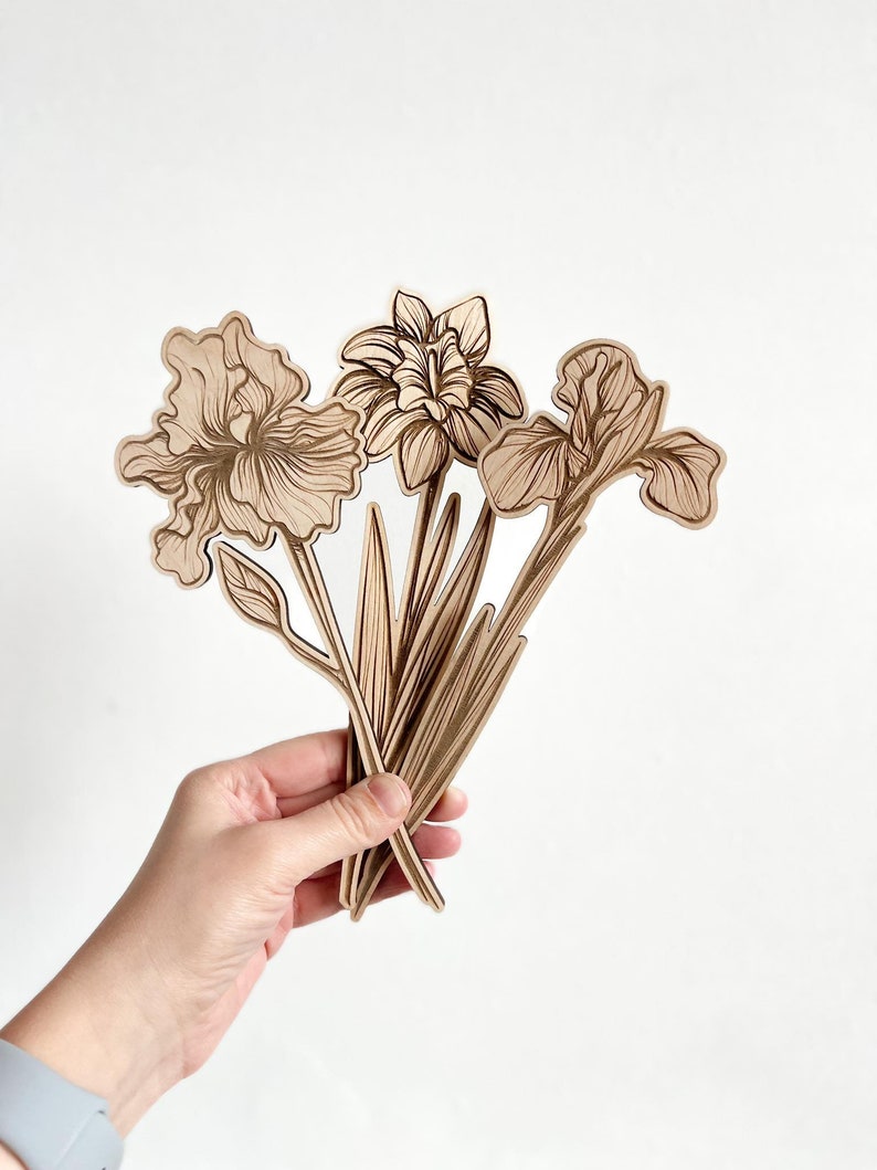 May include: A set of three wooden iris flower cutouts in a light brown color. The flowers are arranged in a bouquet and are held in a hand.