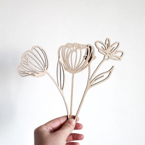 May include: A set of three wooden flower cutouts, each with a unique design. The flowers are in a bouquet shape and are held in a hand.