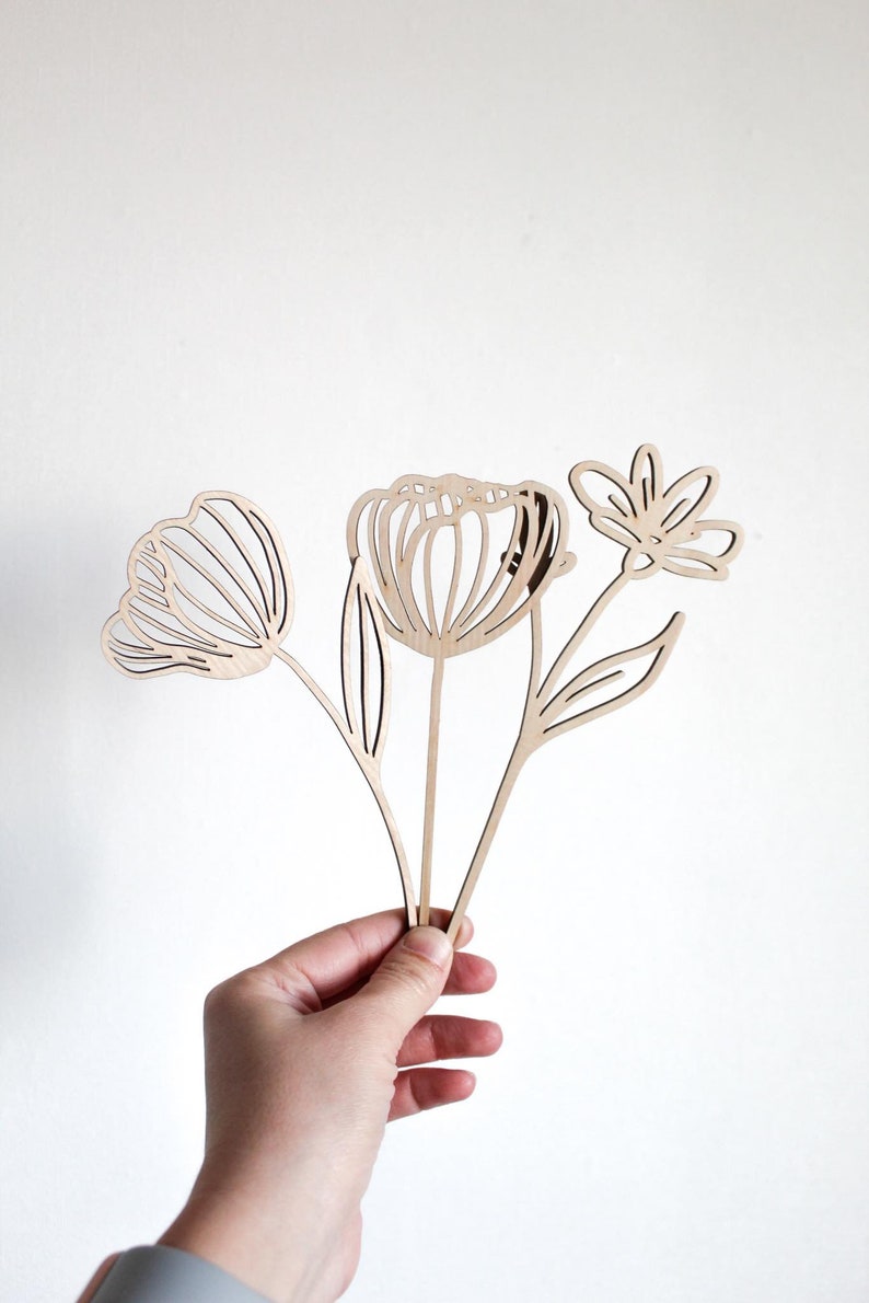 May include: A set of three wooden flower cutouts, each with a unique design. The flowers are in a bouquet shape and are held in a hand.
