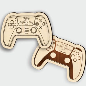 K&ouml;nnte beinhalten: Zwei Holzschilder in Form eines Gamecontrollers. Eines ist hellfarbig mit dem Text "Happy Father's Day 2025 Love, Liam." Das andere ist dunkel gef&auml;rbt mit dem Text "You are The Best Daddy Ever 2025 Love, Liam."