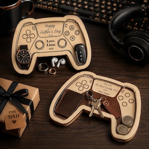 K&ouml;nnte beinhalten: Zwei Holztabletts in Form eines Controllers mit eingravierten Details. Eines enth&auml;lt eine Uhr, Ringe, Ohrh&ouml;rer und einen Autoschl&uuml;ssel. Das andere hat eine Brieftasche, Schl&uuml;ssel und M&uuml;nzen. Der Text lautet "Happy Father's Day 2025" und "You are The Best Daddy Ever 2025".
