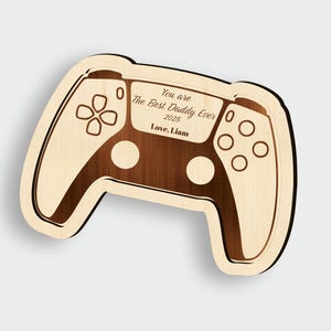 K&ouml;nnte beinhalten: Holzplakette in Form eines Gamecontrollers mit der Aufschrift "You are The Best Daddy Ever 2025 Love, Liam." Das Design verwendet helles Holz mit dunkleren Holzdetails, die einen Spielekonsolen-Controller nachahmen.
