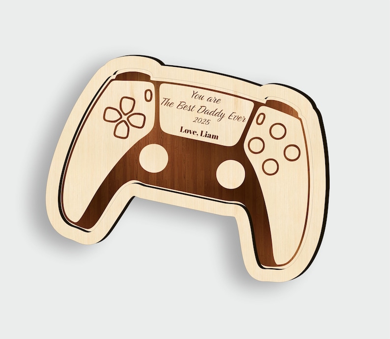 K&ouml;nnte beinhalten: Holzplakette in Form eines Gamecontrollers mit der Aufschrift "You are The Best Daddy Ever 2025 Love, Liam." Das Design verwendet helles Holz mit dunkleren Holzdetails, die einen Spielekonsolen-Controller nachahmen.