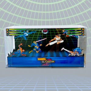 May include: A Marvel vs. Capcom 2 diorama featuring several characters in action poses. The scene is set within a clear display case, with a blue and white background. The game's logo is visible at the bottom.