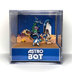 May include: A clear acrylic display box containing a 3D diorama of Astro Bot characters. The front panel features the text ASTRO BOT in white. Inside, a small white and blue robot stands on a tan base before a blue background with printed figures.