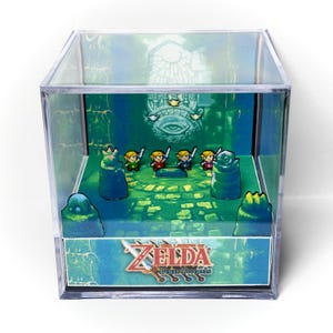 Puede incluir: Un cubo de exhibición de acrílico transparente que contiene un diorama 3D de The Legend of Zelda: Four Swords. Dentro, cuatro pequeñas figuras se encuentran sobre un suelo de piedra verde ante una pared con un emblema de ojo. El logo es visible.