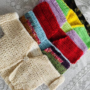 May include: A set of crocheted baby sweaters in various colors, including beige, red, green, blue, purple, and yellow. The sweaters are made with a soft, textured yarn and have a simple, classic design.
