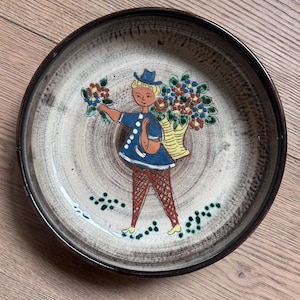 Op de afbeelding: Een ronde keramische schaal met een donkerbruine rand en een beige centrum, voorzien van een handgeschilderde illustratie van een figuur in een blauw jasje en hoed, die een grote mand met kleurrijke bloemen op de rug draagt.