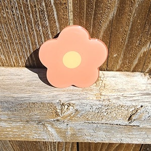May include: A peach-colored flower with a yellow center. The flower is made of plastic and has a glossy finish. It is sitting on a wooden surface.