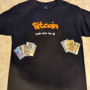 Puede incluir: Una camiseta negra con el texto "Bitcoin Gotta stack 'em all" en naranja. La camiseta tiene una pila de cartas de Pokémon holográficas en la parte delantera.