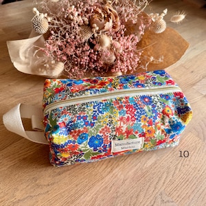 Peut inclure: Une trousse rectangulaire à fermeture éclair avec un imprimé floral et une poignée crème. La trousse présente un motif coloré de fleurs rouges, bleues et jaunes sur fond blanc. La fermeture éclair est dorée et l'étiquette indique "Manufacture Marlou."