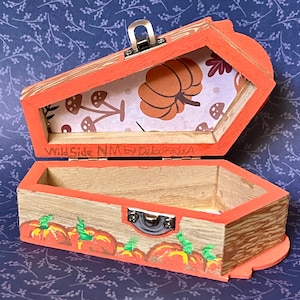 May include: An open, orange coffin-shaped wooden box with a hinged lid. The interior of the lid features a pumpkin and mushroom pattern. The box is decorated with painted pumpkins and the text "Wild Side NM by Deborah A".