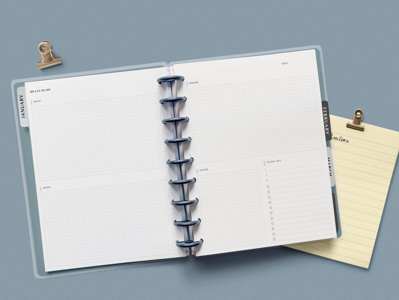 May include: An open planner with a blue cover and black rings. The left page has the word "January" and the right page has the word "February". The pages are blank and have a grid pattern.