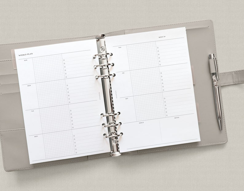 May include: A white weekly planner with a gray binder and a silver pen. The planner has a gridded layout for each day of the week, with space for notes and goals. The planner is open to the week of the 22nd.