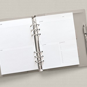 May include: An open gray leather bound planner with a silver binder. The planner has a gridded page with the words "Brain Dump", "Must", "Want", "Date", "Notes", and "To Do List". A silver pen is in the pen loop.