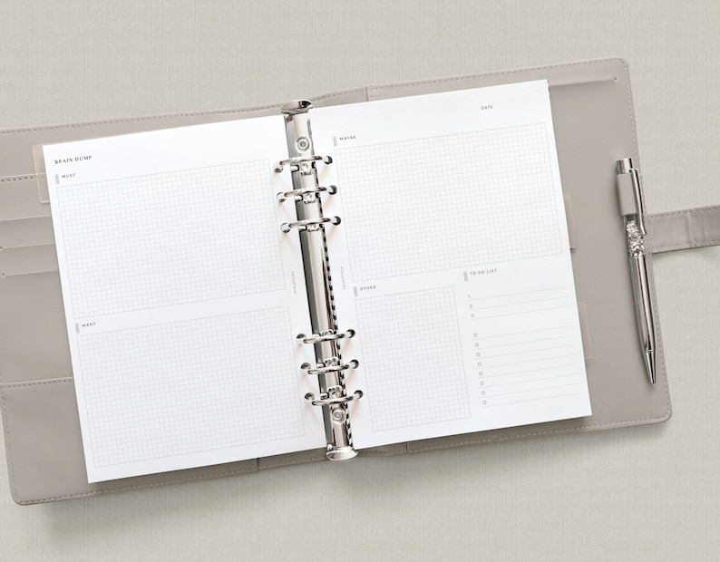 May include: An open gray leather bound planner with a silver binder. The planner has a gridded page with the words "Brain Dump", "Must", "Want", "Date", "Notes", and "To Do List". A silver pen is in the pen loop.