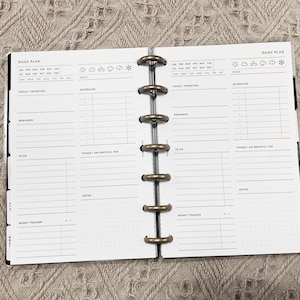May include: A daily planner with a black cover and gold rings. The planner has sections for focus, priorities, schedule, reminders, to do, things I am grateful for, notes, and money tracker. The planner also has a section for mood with icons for sun, clouds, rain, and snow.