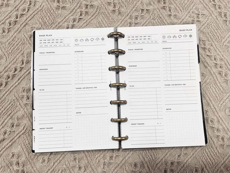 May include: A daily planner with a black cover and gold rings. The planner has sections for focus, priorities, schedule, reminders, to do, things I am grateful for, notes, and money tracker. The planner also has a section for mood with icons for sun, clouds, rain, and snow.