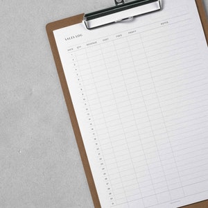 以下が含まれることがあります： 銀色のクリップが付いた茶色のクリップボードに白い売上ログ。ログには、日付、数量、収益、費用、手数料、利益、メモの列があります。