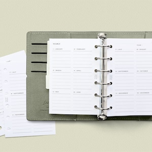 K&ouml;nnte beinhalten: Ein grauer, ledergebundener Taschenplaner mit einem j&auml;hrlichen Kalendereinsatz. Der Kalender ist in 12 Monate unterteilt, mit Zeilen zum Schreiben von Notizen.