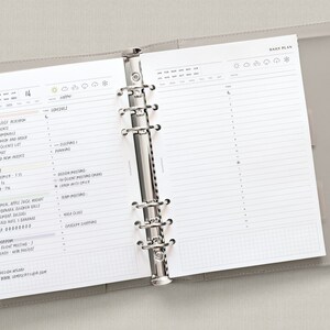 Puede incluir: Una agenda diaria con una cubierta gris y anillos plateados. La agenda est&aacute; abierta en una p&aacute;gina con un plan diario para el 14. La p&aacute;gina tiene una secci&oacute;n para "Get it Done", "Expenses", "Meals", "Water", "Tomorrow" y "Design Affair". La p&aacute;gina tambi&eacute;n tiene una secci&oacute;n para "Time" con una cuadr&iacute;cula para la planificaci&oacute;n.