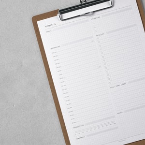 K&ouml;nnte beinhalten: Ein braunes Clipboard mit einem wei&szlig;en Blatt Papier. Das Papier hat ein Tagesplaner-Layout mit Abschnitten f&uuml;r Priorit&auml;ten, To-Do-Liste, Zeitplan, Anrufe/E-Mails/SMS, Notizen und Produktivit&auml;t.