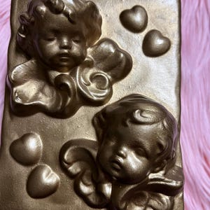 May include: A gold-colored rectangular plaque featuring two cherubic figures with wings and three heart-shaped accents. The cherubs are in a bas-relief style, with detailed facial features and curly hair. The plaque has a textured surface.