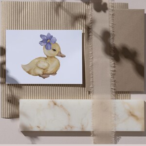 May include: A watercolor illustration of a yellow duckling wearing a purple flower on its head. The duckling is on a white card, surrounded by neutral-toned stationery and a marble-like block.