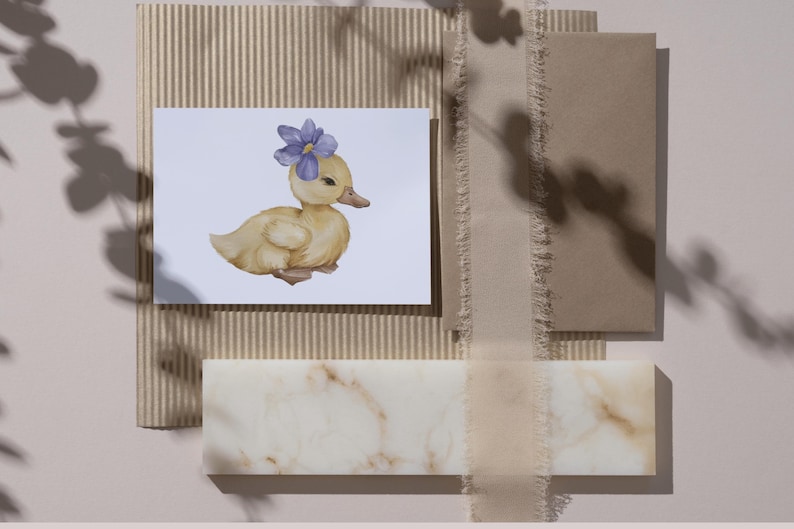 May include: A watercolor illustration of a yellow duckling wearing a purple flower on its head. The duckling is on a white card, surrounded by neutral-toned stationery and a marble-like block.