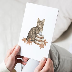 May include: A greeting card featuring a watercolor illustration of a tabby cat sitting next to a layered cake with a single candle. The card is decorated with autumn leaves and has a white background.