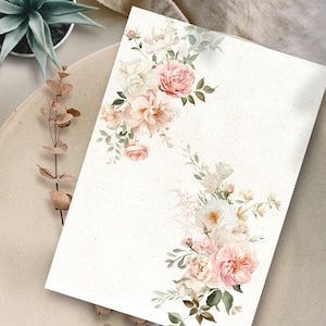 May include: A blank white card with a floral border. The border is made up of pink and white roses, green leaves, and white flowers. The card is on a beige surface.