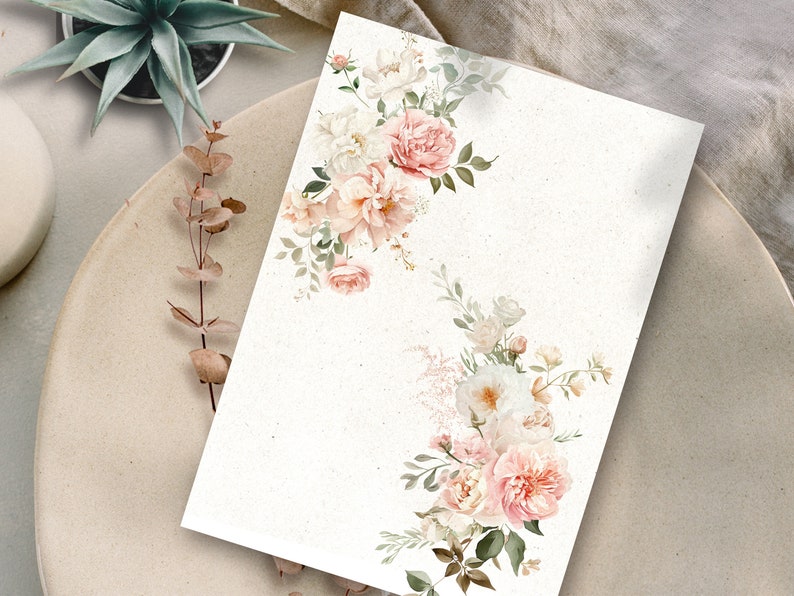 May include: A blank white card with a floral border. The border is made up of pink and white roses, green leaves, and white flowers. The card is on a beige surface.