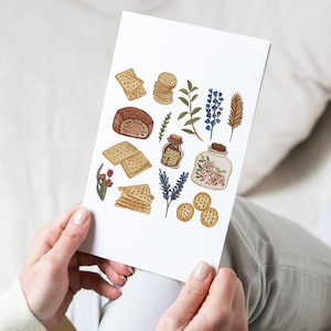 May include: A white card featuring watercolor illustrations of food items and botanicals. The image includes crackers, bread, herbs, and jars filled with spices and flowers. The color palette is soft, with warm browns, greens, and blues. The card is held by a person.