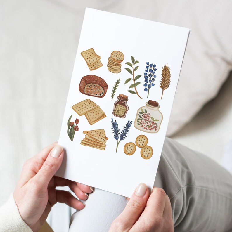 May include: A white card featuring watercolor illustrations of food items and botanicals. The image includes crackers, bread, herbs, and jars filled with spices and flowers. The color palette is soft, with warm browns, greens, and blues. The card is held by a person.