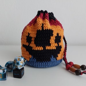 May include: A crocheted dice bag with a black paw print design. The bag is red, orange, and blue. It has a drawstring closure and wooden beads. There are five dice on the table, two are blue and white with gold numbers, two are black with white numbers, and one is black with gold numbers.