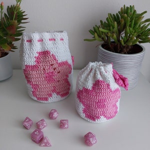 May include: Two crocheted drawstring bags with pink flower designs. The bags are made with white and pink yarn. There are several pink dice on a white surface.