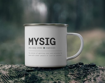 Könnte beinhalten: Weißer Emaille-Becher mit dem Wort "MYSIG" in Schwarz aufgedruckt. Der Becher enthält auch die Definition des Wortes, ein schwedisches Verb. Der Becher steht auf einem Baumstamm.