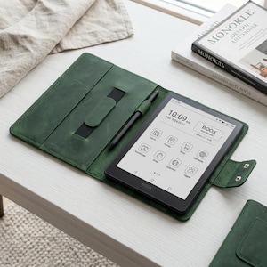 Puede incluir: Una funda de cuero verde para lector electrónico con un lápiz óptico negro y un lector electrónico digital que muestra la hora y la fecha. El lector electrónico está abierto, mostrando iconos de aplicaciones. La funda está sobre una superficie blanca.