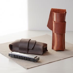 Puede incluir: Una armónica con acabado negro y plateado, junto a dos estuches de cuero. Un estuche es de color marrón oscuro, de diseño rectangular con cierre de solapa. El otro es un estuche vertical marrón claro.