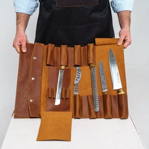 May include: A brown leather knife roll with several knives of varying sizes. The roll is open, displaying the knives in individual slots. One knife has a serrated edge, and another has the word "PIZZA" cut into the blade. The roll is held open by a person.