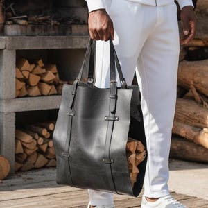 May include: A black leather tote bag with two handles and adjustable straps. The bag has a rectangular shape and is held by a person wearing a white tracksuit and white sneakers. The background includes a brick structure and a pile of firewood.